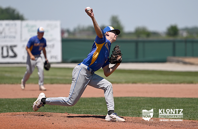 Baseball pitcher in blue and yellow uniform throws a pitch from the mound while an infielder watches in the background.
