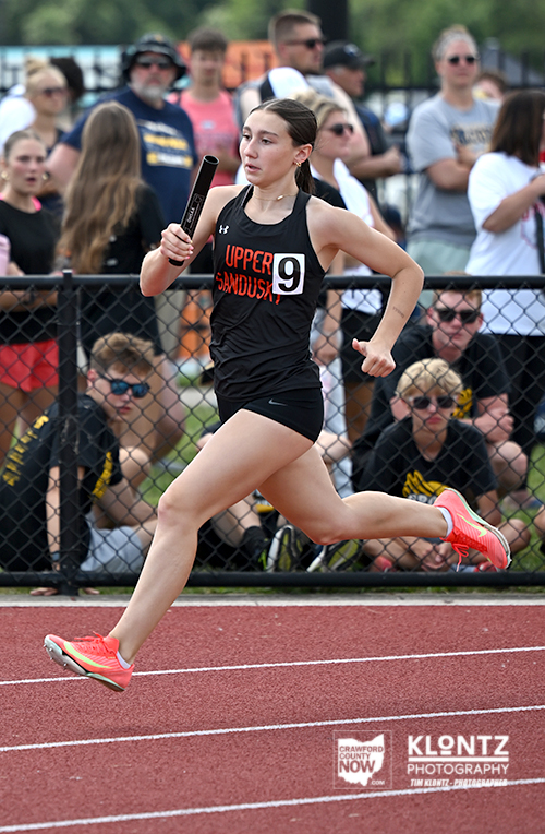 Female sprinter in a black uniform holding a baton, mid-race on a red track with spectators behind a chain-link fence.