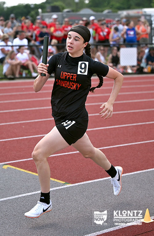 Female baton sprinter in black Upper Sandusky uniform (number 9) runs on a red track, holding a relay baton with spectators in the background.