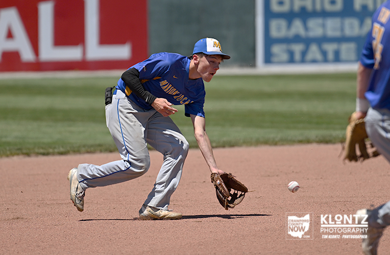 Baseball infielder in a blue jersey reaches for a ground ball with his glove on a dirt infield as a ball rolls toward him.