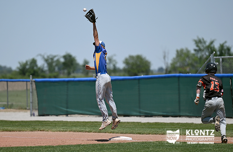 Baseball player in blue and yellow jumps to catch a rising ball at first base as a runner in black approaches on a sunny day.
