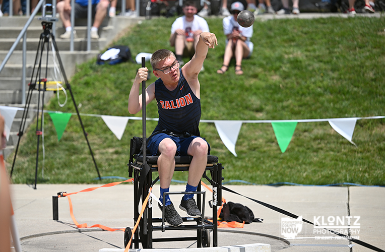 Male para-athlete in a sports wheelchair throwing a shot put at an outdoor track meet, with spectators watching from a grassy slope and bunting in the background.