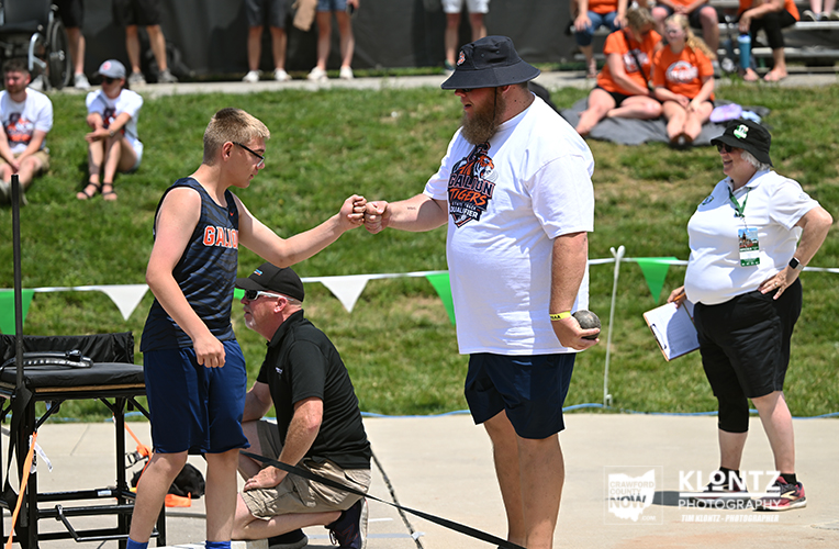 Two participants fist-bump on a sunny field during a youth sports event, with spectators watching on a grassy hill behind them.