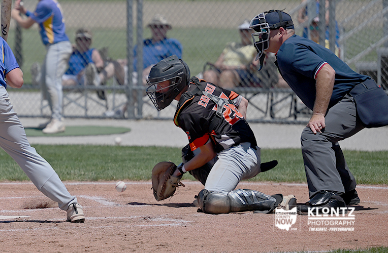 Baseball catcher in black/orange crouches at home plate with glove ready as a ball rolls in; umpire stands behind.