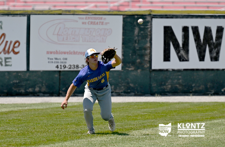Female baseball player in a blue jersey and gray pants reaches with her glove to catch a ball in the outfield; advertising boards line the wall behind her.