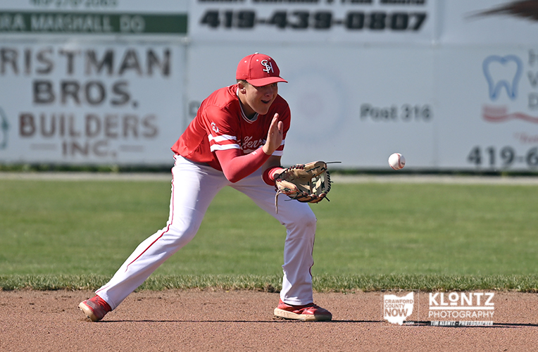 Baseball player in a red uniform fields a ground ball with glove low, preparing to throw on a dirt infield.