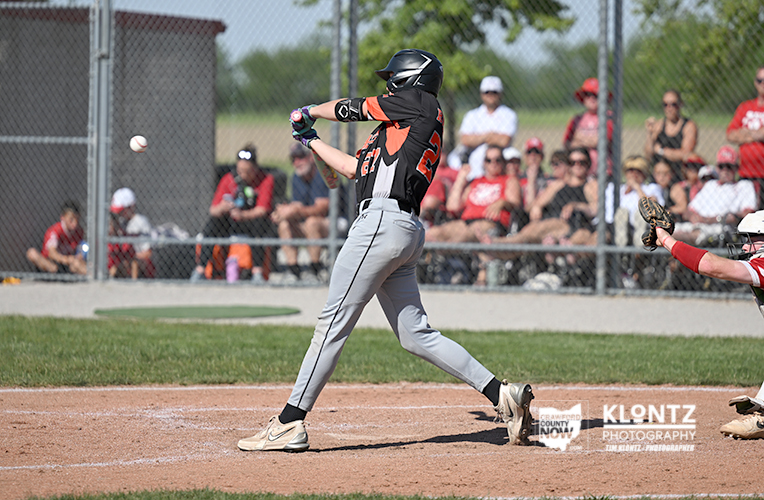 Baseball batter in black-and-orange uniform swings at a pitch on the field with spectators behind a chain-link fence.