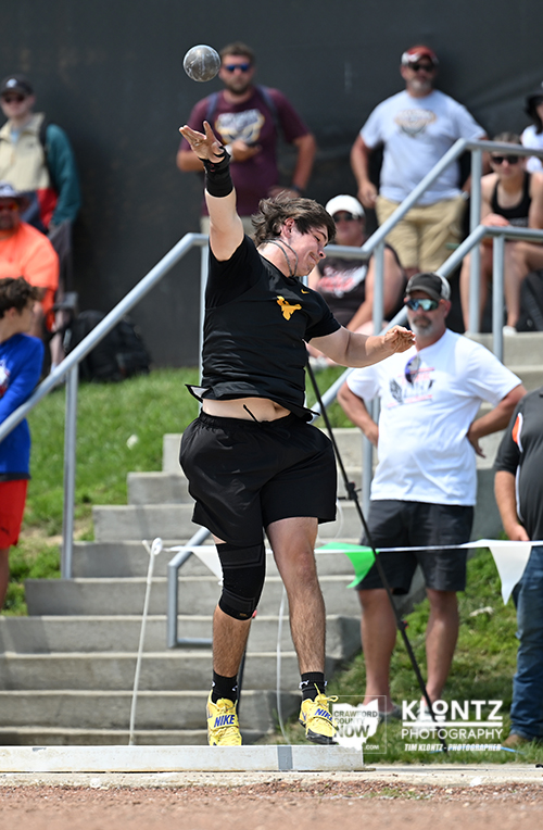 Male athlete mid-throw in a black outfit, launching a discus during an outdoor track event with spectators on bleachers in the background.
