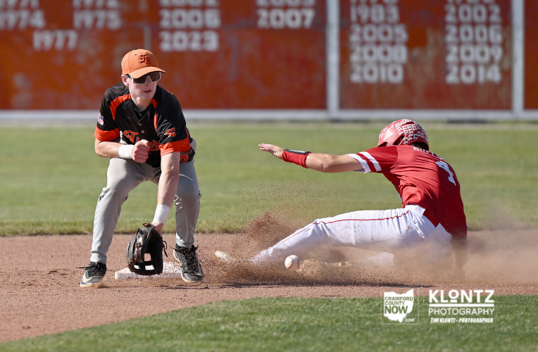 A baseball fielder in orange and black crouches with a glove as a baserunner slides into a base, dust kicking up from the dirt.