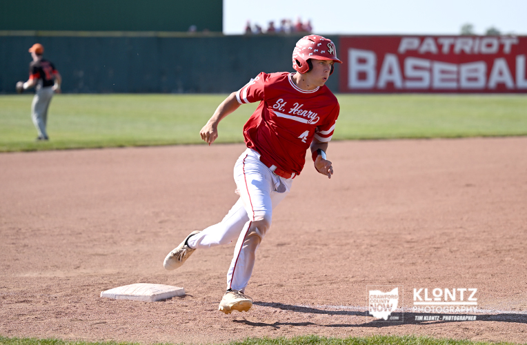 Baseball player in a red uniform and helmet sprinting toward first base on a dirt infield, with a 'Patriot Baseball' banner in the background.