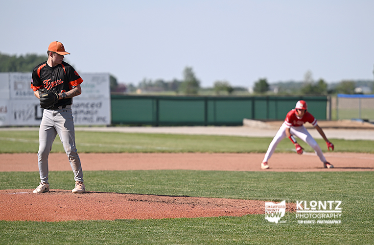 Baseball scene: pitcher on the mound in black-and-orange uniform prepares to throw, while a fielder in red stands near a base on a sunny day.