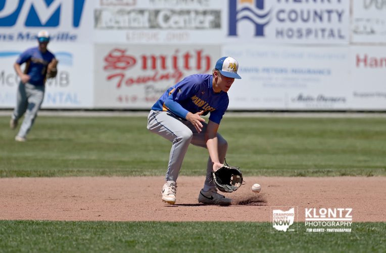 Youth baseball player in blue jersey and gray pants crouches to field a ground ball, glove low near the dirt.