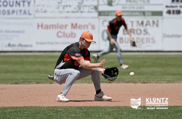 Baseball fielder in an orange cap and black uniform crouches to field a ground ball with his glove near the infield dirt, ads on the outfield wall in the background.