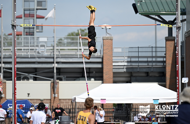 Pole vaulter in mid-air clearing the bar at a track meet, holding the pole with a stadium backdrop and spectators nearby