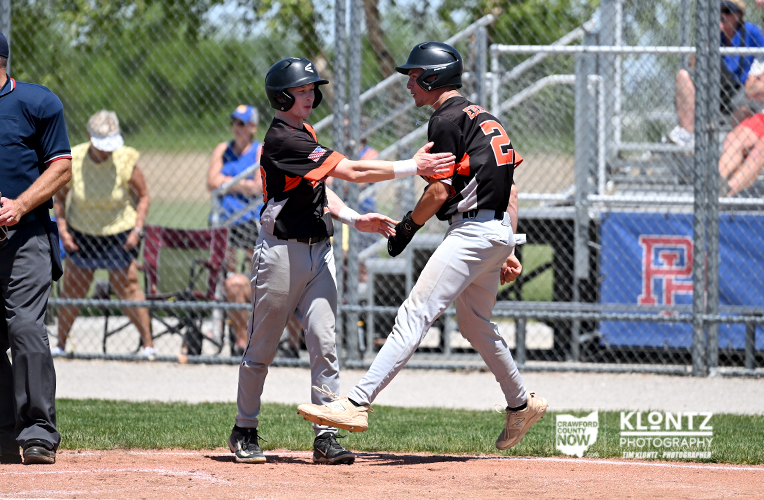 Two young baseball players in black and orange uniforms high-five on the infield near a baserunner, wearing helmets.
