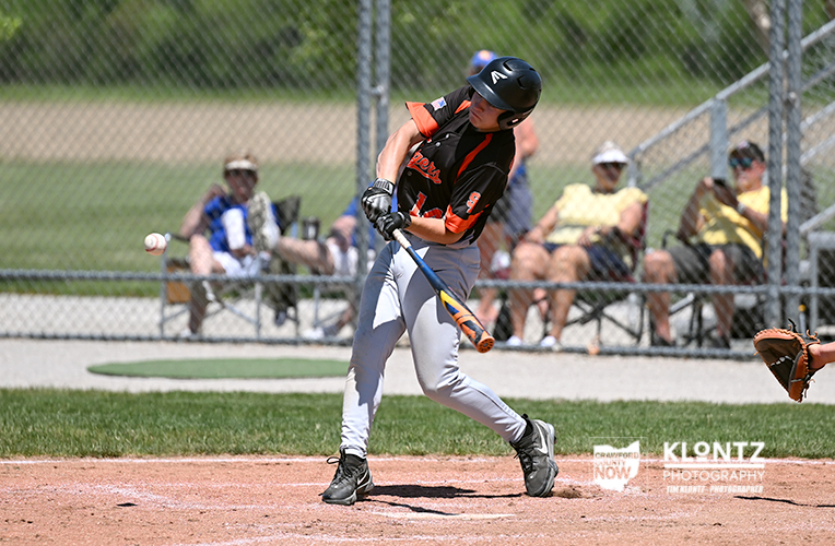 Baseball batter in black and orange swings at a pitch on a dirt infield with spectators behind a chain-link fence.