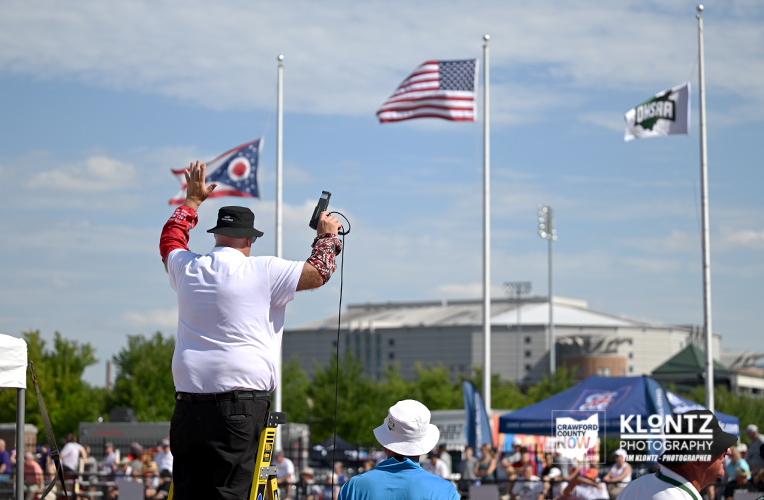 Speaker on a yellow ladder addressing a crowd at an outdoor event, raising his hand while holding a microphone, with flags and tents in the background.