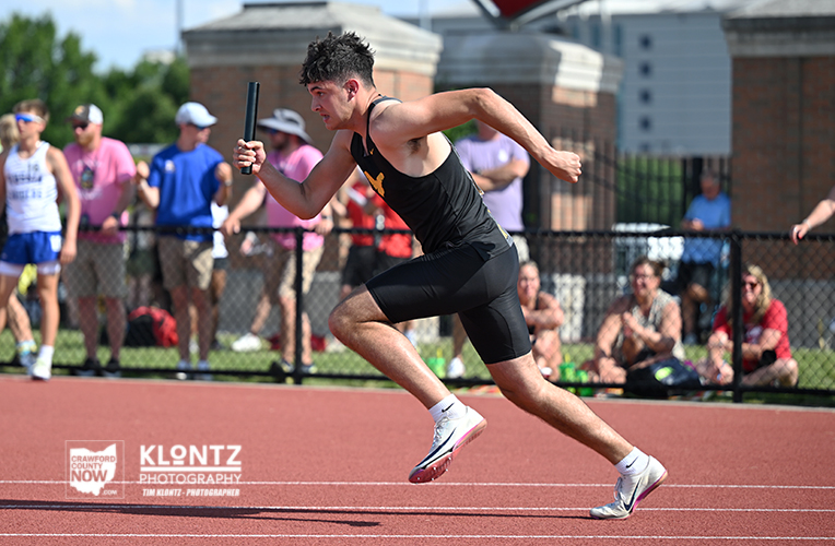 Male sprinter in a black uniform runs on a red track, holding a baton amid cheering spectators behind a fence.