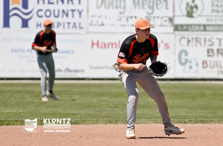 Baseball player in an orange cap and black-orange uniform crouches on the dirt pitcher's mound with glove ready as a teammate stands in the grass in the background.