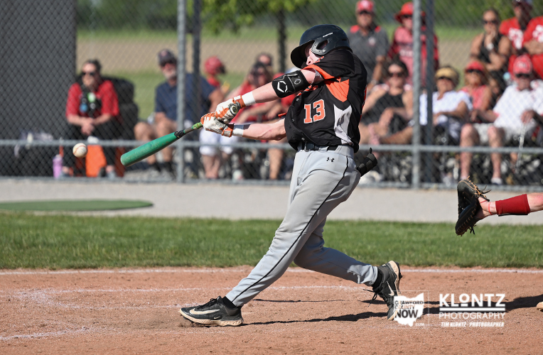 Baseball batter in black/orange swings at a pitch with a green bat, crowd watching behind a chain-link fence.