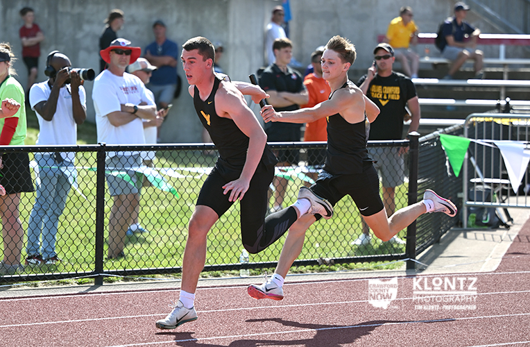 Two male sprinters in black uniforms race on a track; one reaches out to the other during a relay handoff as spectators watch behind a fence.