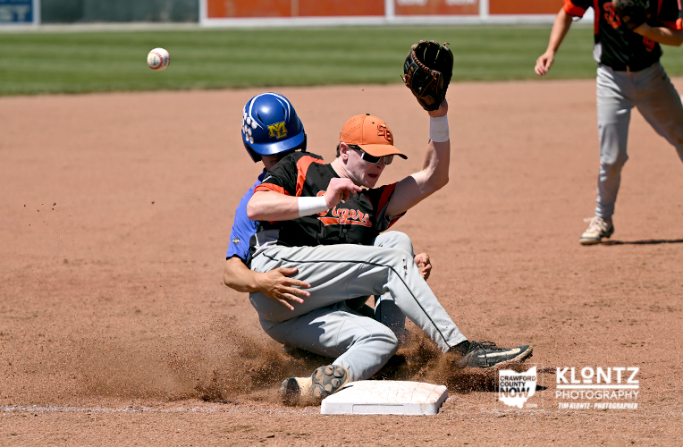 Runner slides into base while fielder reaches with glove to tag; ball is in the air above the dirt infield as dust rises.