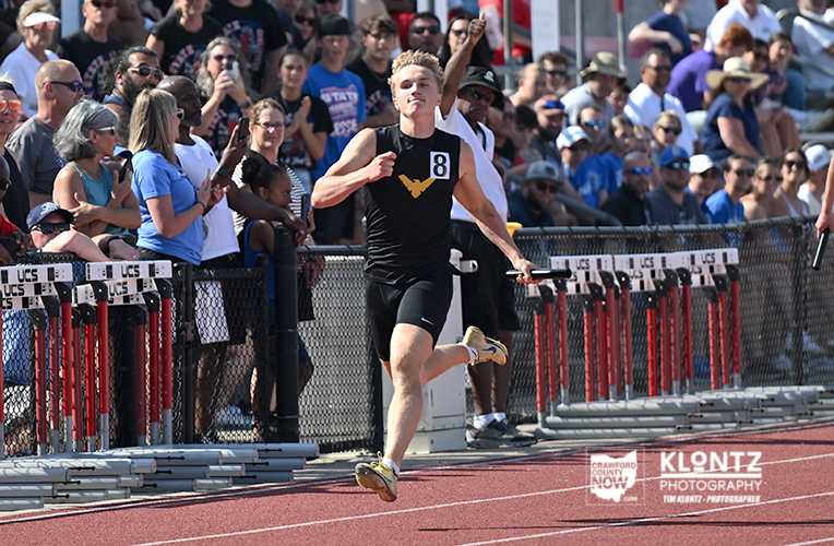 Male sprinter in black kit (bib 8) crossing the finish line on a red track, crowd cheering behind the fence.
