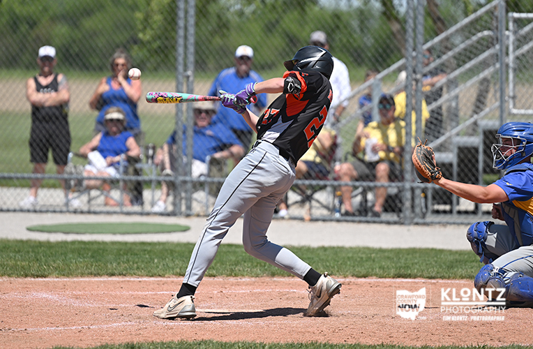 Baseball batter in black and red swings at a pitch near home plate, with a blue-caught catcher reaching to receive the ball's path behind him.