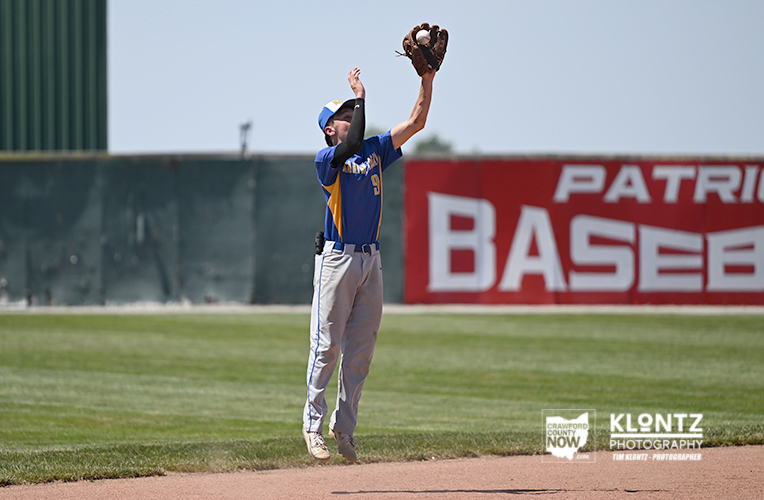 Baseball player in blue-yellow uniform reaches up to catch a ball on a grassy field near a red sign that reads BASEB... (stadium background)