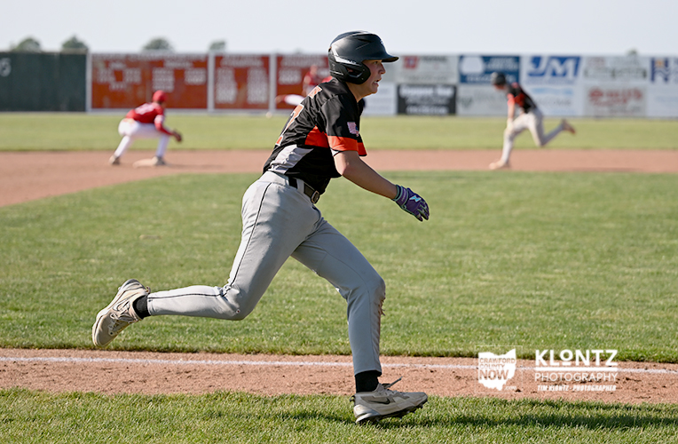 Baseball player in black-and-red uniform running on a dirt basepath, helmet on, during a game with a pitcher in the background.