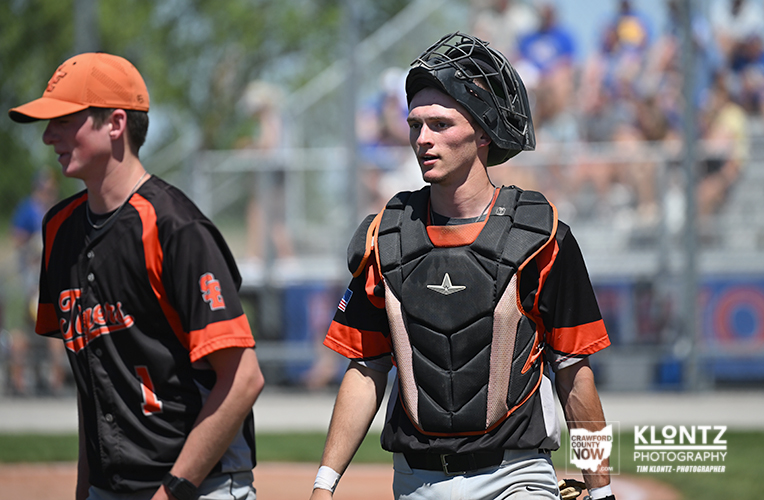 Two baseball players on a field; one in catcher's gear walking beside a teammate in black-orange uniform.