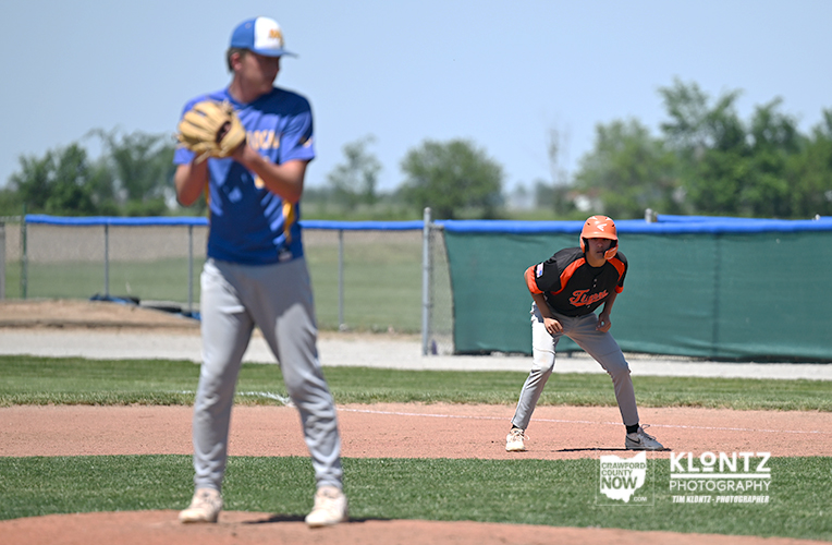 Blue-uniform pitcher in the foreground winds up on the mound while a baserunner in an orange helmet waits on the infield.