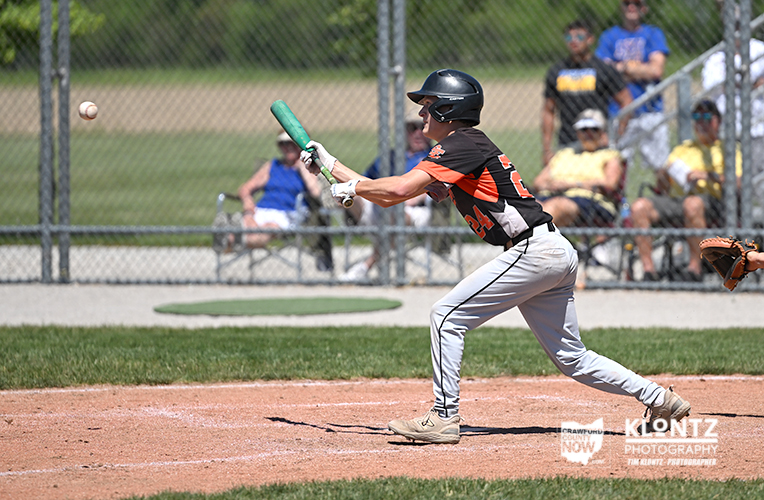 Baseball batter in black and orange uniform swings a green bat at a pitched ball on a sunny field with a chain-link fence and spectators in the background.