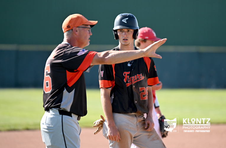 Coach in black and orange uniform (number 6) signals with his arm raised to a young catcher/runner during a baseball game on the infield.