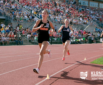 Three female sprinters race on a red track as a large crowd watches from stadium stands.