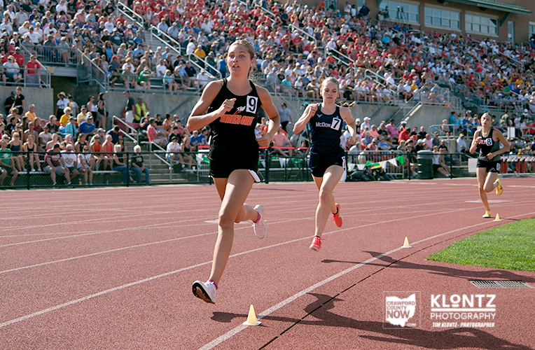 Three female sprinters race on a red track as a large crowd watches from stadium stands.