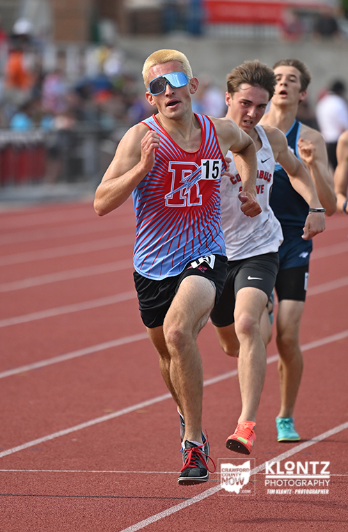 Male sprinters race on a red track; lead runner wears a blue-red striped jersey with bib 15 and sunglasses, mid-stride ahead of rivals.
