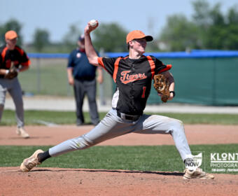 Right-handed pitcher in a black and orange Tigers uniform delivers a pitch from the mound, with teammates and umpire in the background.