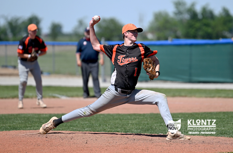 Right-handed pitcher in a black and orange Tigers uniform delivers a pitch from the mound, with teammates and umpire in the background.
