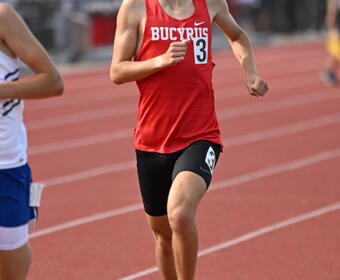Male sprinter in a red Bucyrus jersey and black shorts runs on a red track during a race, number 3 visible on his chest.