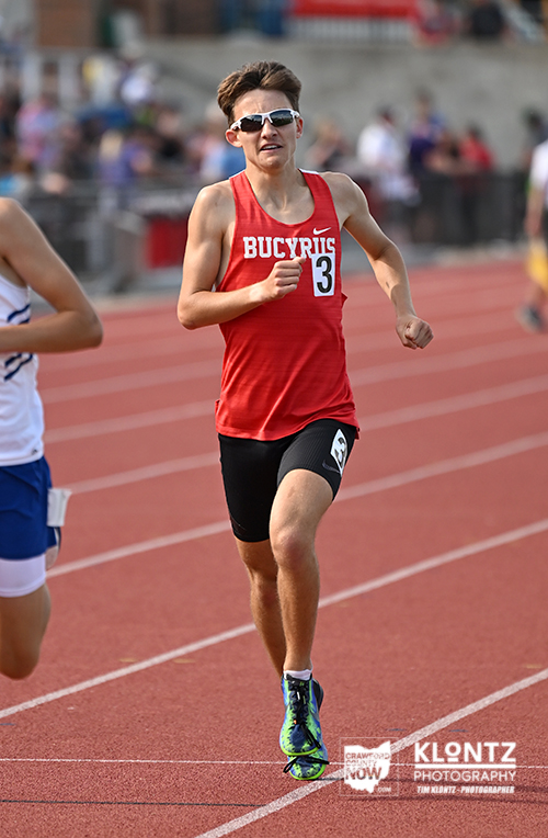 Male sprinter in a red Bucyrus jersey and black shorts runs on a red track during a race, number 3 visible on his chest.