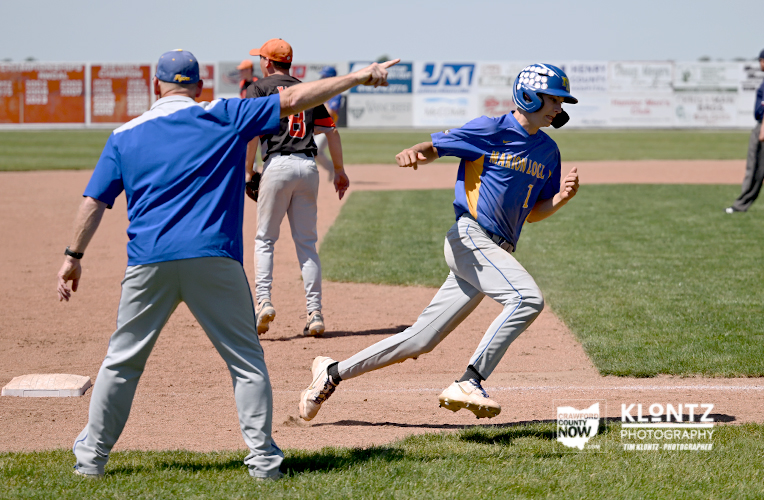 Baseball action: a blue-uniform runner slides into a base while a coach points and an orange-uniform fielder stands nearby on the infield.