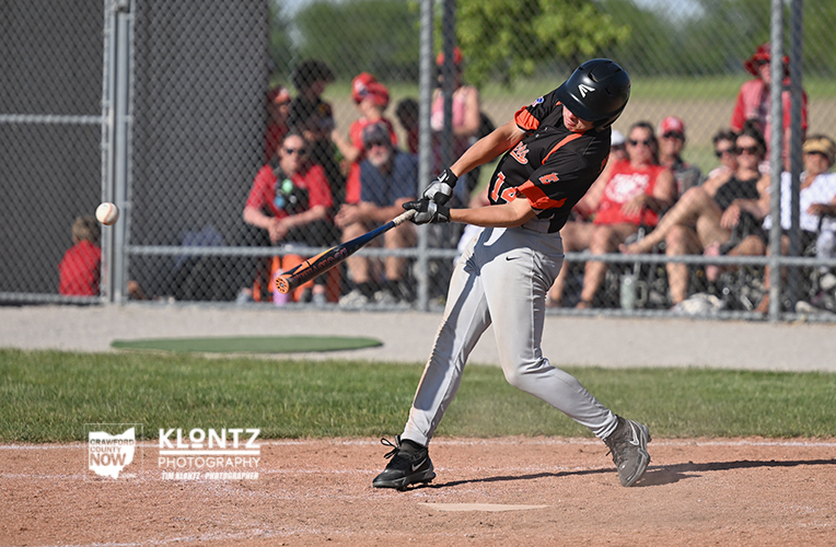 Young baseball player in a black and orange uniform swings at a pitch during a game, with a chain-link fence and spectators in the background.