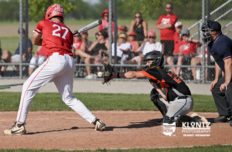 Baseball batter in red swings at a pitch as the catcher in black-orange dives to field the ball near home plate.