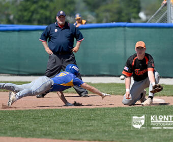 Baseball action: a blue-and-yellow uniformed runner slides headfirst into a base while an orange-and-black fielder kneels nearby and an umpire watches in the background.
