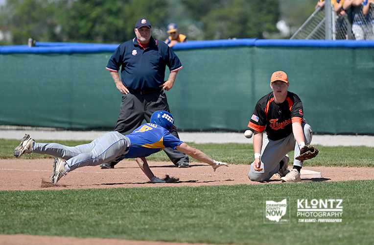 Baseball action: a blue-and-yellow uniformed runner slides headfirst into a base while an orange-and-black fielder kneels nearby and an umpire watches in the background.