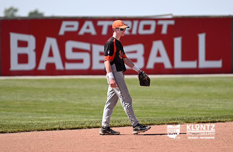 Young baseball player in orange and black uniform walking on the infield with a glove, against a red 'PATRIOT BASEBALL' banner in the background.