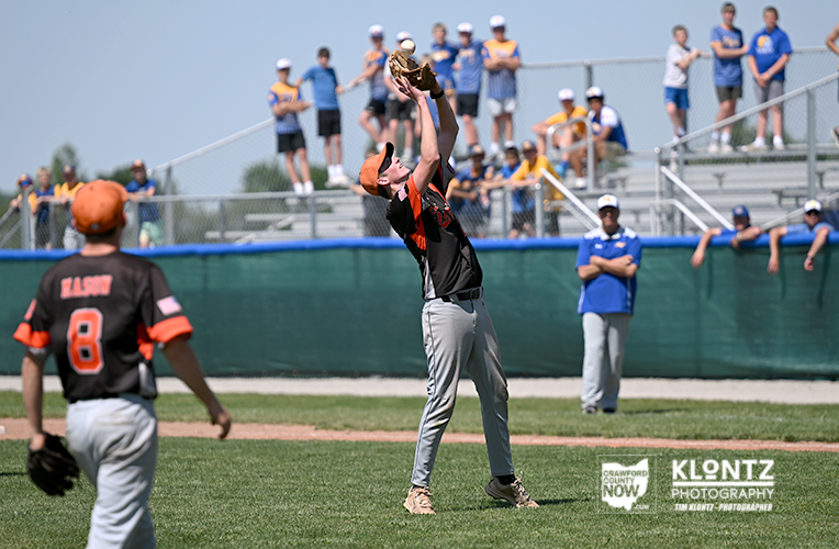Boy in black-orange jersey reaches up to catch a ball during a baseball game as spectators watch from the stands.