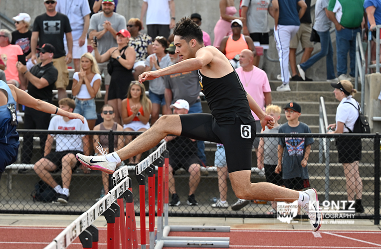 Hurdler in black athletic gear clears a hurdle during a track race, with a cheering crowd in the stands behind him.