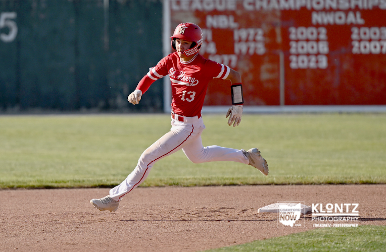 Baseball player in a red uniform sprinting on the infield dirt with a green outfield and scoreboard in the background.